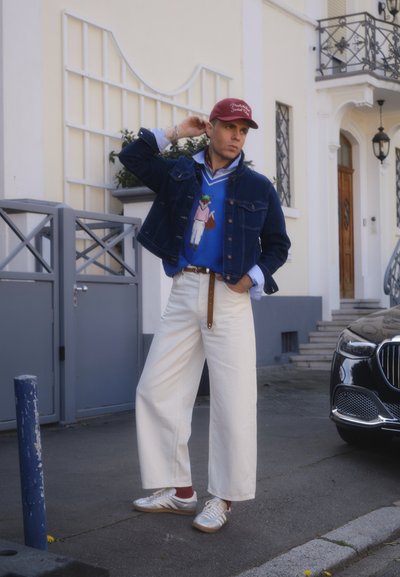 Hombre con pantalones anchos blancos, suéter azul con gráfico de oso, chaqueta vaquera, gorra roja y zapatillas, de pie en la calle cerca de un coche negro y un edificio.