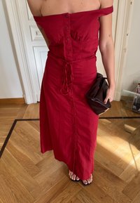 Czerwona sukienka z odsłoniętymi ramionami, dopasowanym gorsetem, zapinana na guziki z przodu, z paskiem wciętym w talii. Dodatek: brązowa torebka kopertówka. Płaskie sandały. Tło z drewnianą podłogą.