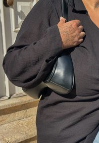 Sac en cuir noir porté sous le bras, présentant une texture lisse et une forme arrondie. La tenue comprend une chemise noire ample à manches longues avec des détails subtils.