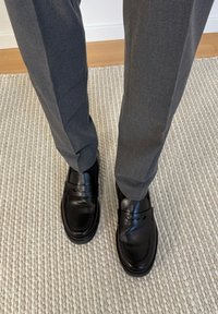Personne portant un pantalon gris et des mocassins en cuir noir, debout sur un tapis beige texturé, avec un sol en bois en arrière-plan.
