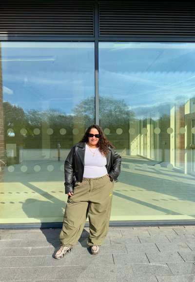 Persona con pelo rizado que lleva gafas de sol, chaqueta de cuero negra, camiseta blanca y pantalones holgados de color oliva, de pie sobre un suelo pavimentado frente a puertas de vidrio.