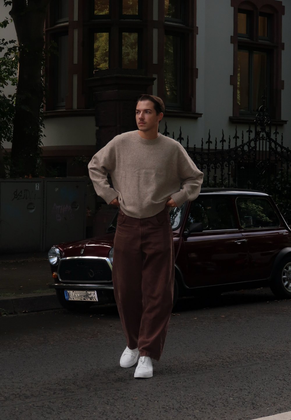 Beige strikket sweater, brede brune fløjlshorts og hvide sneakers. Model står på gaden med en vintage mørkerød bil i baggrunden.