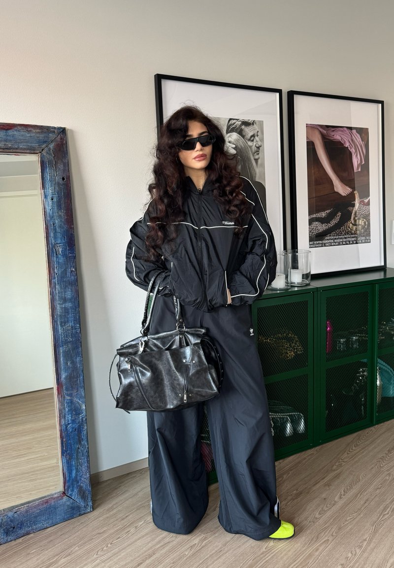 Femme aux cheveux longs et bouclés portant un survêtement noir, des lunettes de soleil foncées, des chaussures jaune néon, et tenant un grand sac en cuir noir à l'intérieur.