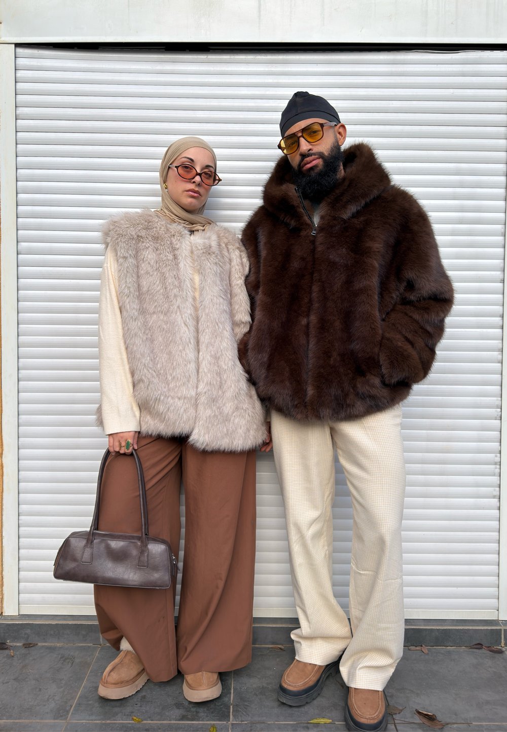Due persone posano davanti a una persiana bianca. Una indossa un gilet beige chiaro in pelliccia sintetica, mentre l'altra indossa una giacca di pelliccia marrone scuro. Entrambe indossano occhiali da sole.
