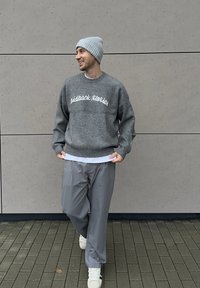 Grauer Pullover mit Aufdruck "laidback stories", kombiniert mit locker sitzender grauer Hose, weißem Unterhemd und hellgrauer, gerippter Mütze. Weiße Turnschuhe.