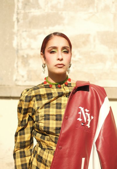 Mujer con sombra de ojos verde y pendientes en forma de corazón, vistiendo un vestido a cuadros amarillos y sosteniendo una chaqueta de cuero roja y blanca de NY sobre su hombro.