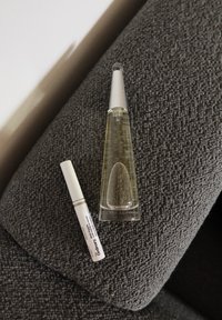 Glasflasche mit einer schmalen Spitze, gefüllt mit klarem Flüssigkeit, neben einem weißen Rohr mit Text. Beide liegen auf einer strukturierten grauen Oberfläche.