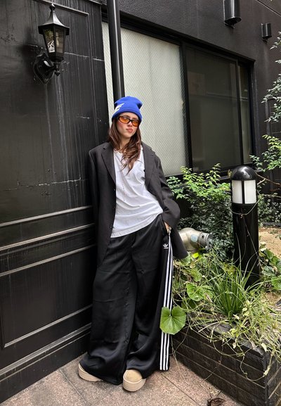 Blazer negro de gran tamaño sobre una camisa blanca, combinado con pantalones deportivos negros con rayas laterales blancas y zapatos slip-on beige, adornado con una boina azul y gafas de sol naranjas.