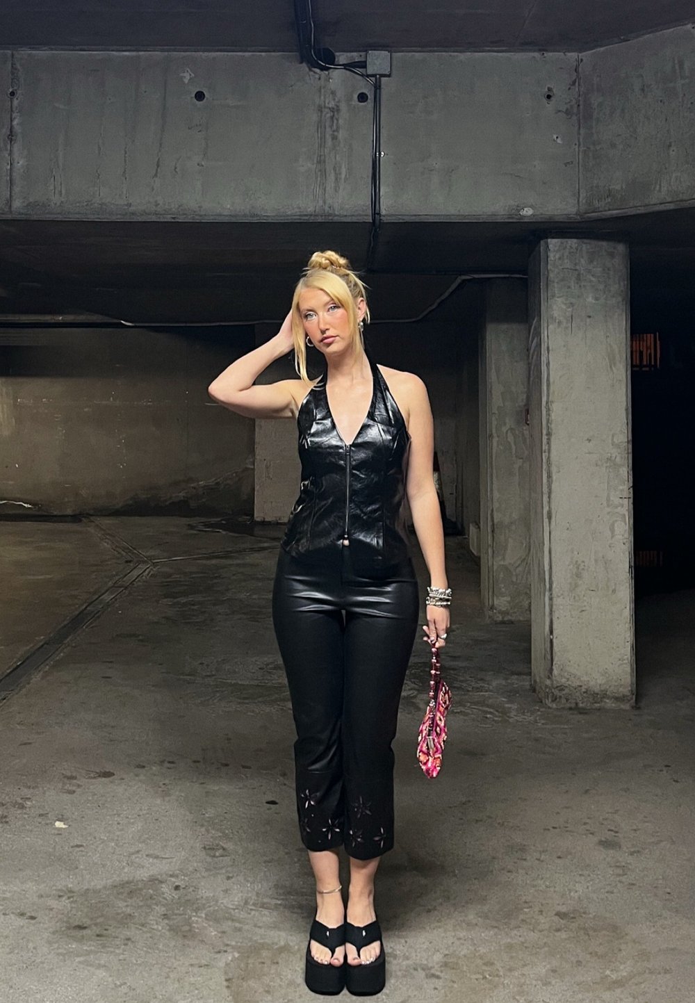 Top halter din piele ecologică neagră, pantaloni negri ajustați cu decupaje și sandale cu platformă. Poartă o geantă roz cu un model. Spațiu interior.