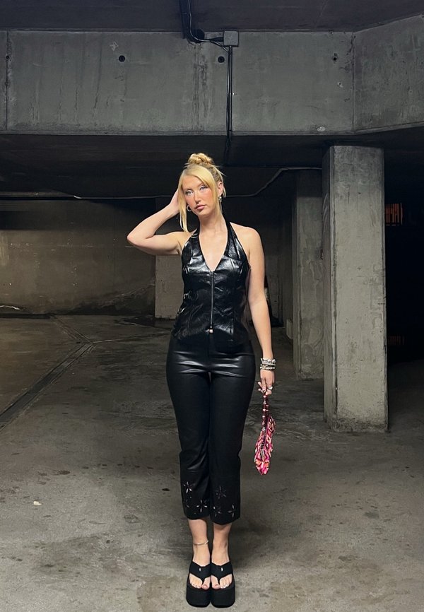 Top halter din piele ecologică neagră, pantaloni negri ajustați cu decupaje și sandale cu platformă. Poartă o geantă roz cu un model. Spațiu interior.