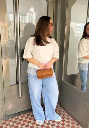 Mujer con camisa blanca de gran tamaño, vaqueros anchos azul claro, zapatillas y bolso marrón, de pie junto a una puerta metálica con espejo.