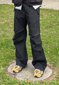 Pantaloni neri larghi con una texture liscia e grandi tasche. Abbinati a scarpe sportive arancioni e grigie con un design a fantasia.
