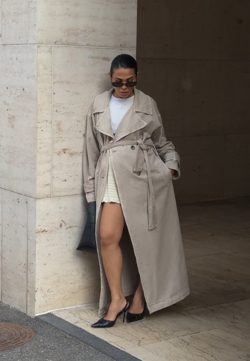 Manteau beige avec col oversize, taille cintrée et manches longues, associé à un haut blanc, un sac à main noir et des escarpins noirs.