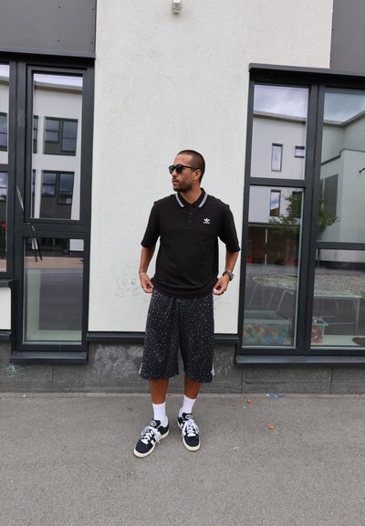 Camisa polo negra con cuello blanco, pantalones cortos negros con estampado, zapatillas negras y calcetines blancos; posando frente a un edificio moderno.