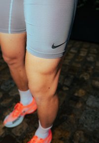 Shorts de sport gris avec un logo Nike noir, fabriqués dans un tissu lisse et extensible, associés à des baskets orange vif et blanches.