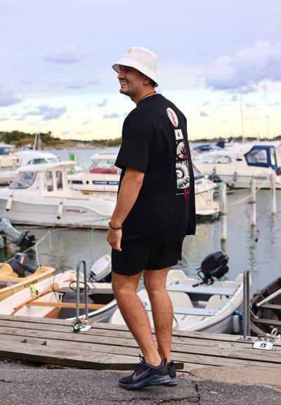 Camiseta negra con estampados gráficos en la parte de atrás, pantalones cortos negros y un sombrero de cubo blanco. Zapatos negros. Agua y barcos al fondo.