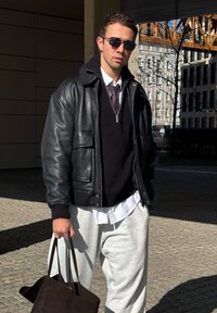 Giacca bomber in pelle nera sopra un maglione scuro, camicia chiara con colletto, pantaloni grigi e porta una borsa a tracolla scura. Occhiali da sole indossati.