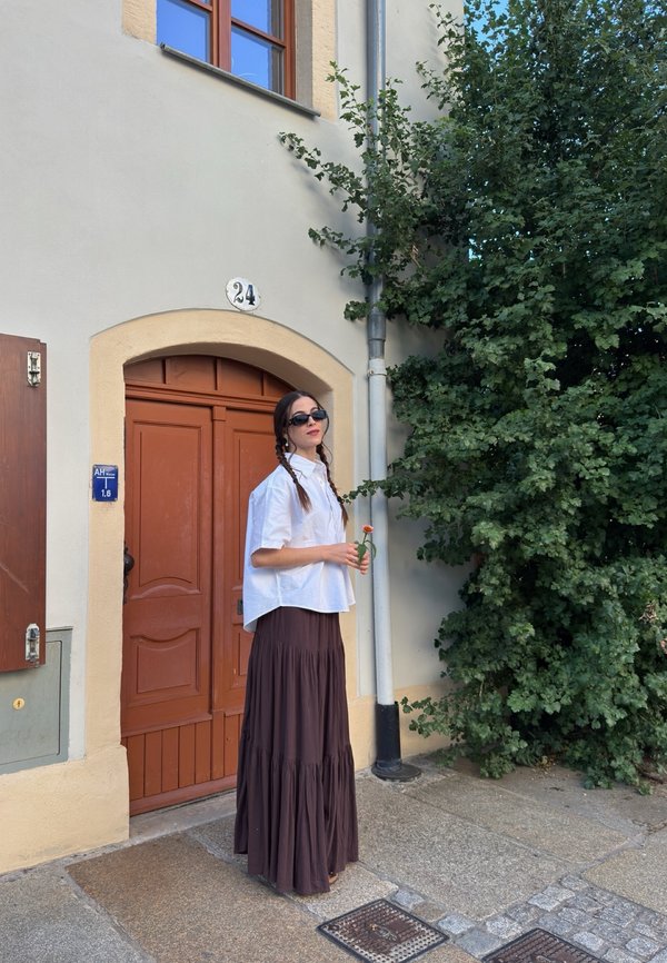 Femme aux cheveux tressés portant des lunettes de soleil, un chemisier blanc et une longue jupe sombre, debout près d'une porte marron tenant une fleur orange près de buissons verts.