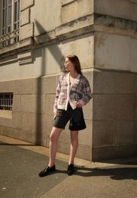 Sweter w różowo-kremową kratę, zapięty, w połączeniu z czarnymi dżinsowymi szortami. Modelka nosi czarne mokasyny i trzyma telefon, stojąc przy teksturowanej ścianie.