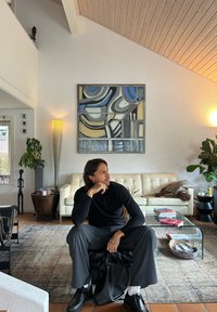 A black textured handbag rests on a seated figure dressed in a black long-sleeve shirt and grey striped trousers, with a cream sofa and abstract wall art in the background.