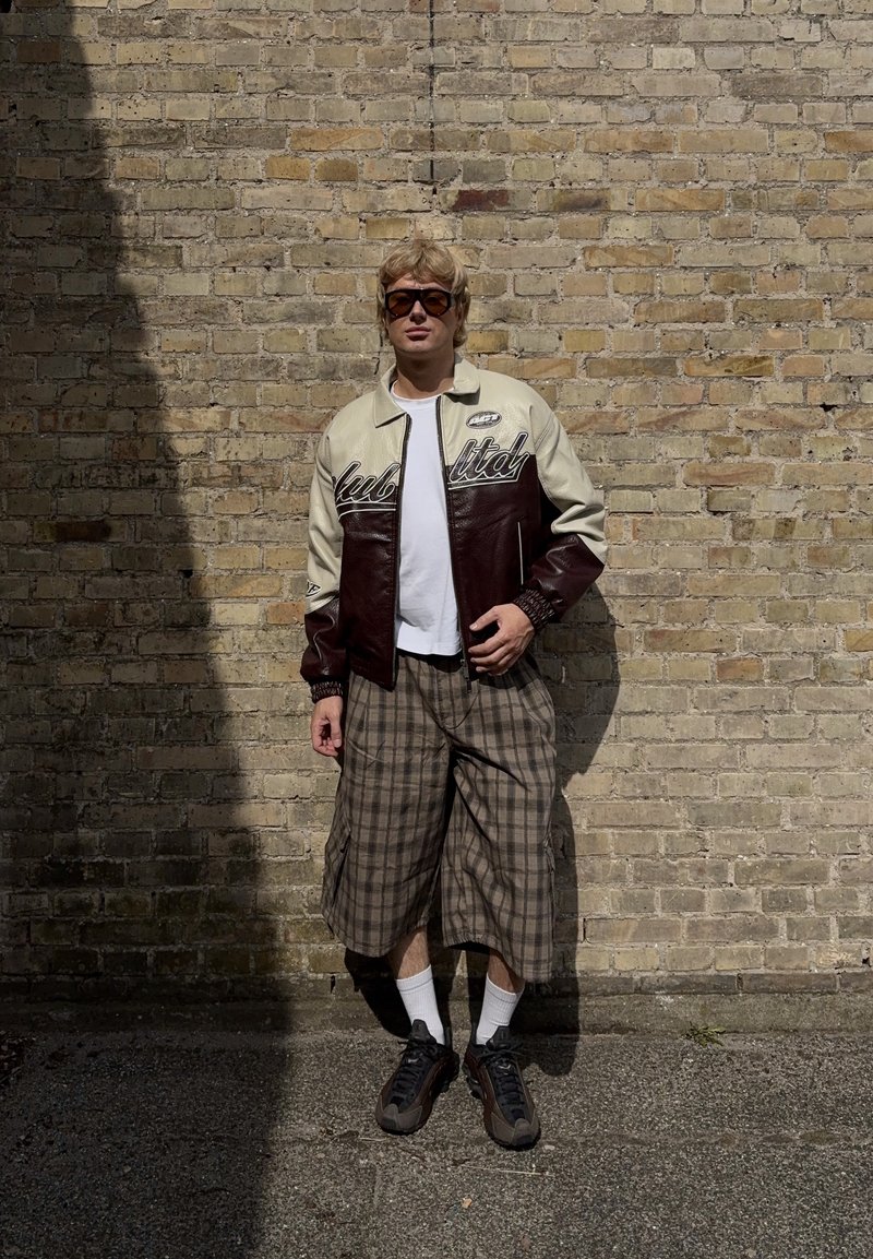 Ξανθός άνθρωπος με υπερμεγέθη γυαλιά ηλίου, δερμάτινο μπουφάν διχρωμίας, λευκό πουκάμισο, καρό φαρδιά σορτς, λευκές κάλτσες και σκούρα αθλητικά παπούτσια, στέκεται δίπλα σε τοίχο από τούβλα.