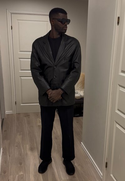 Hombre con gafas de sol negras, blazer de cuero negro, camisa negra, pantalones negros, de pie en el interior cerca de puertas blancas sobre suelo de madera.