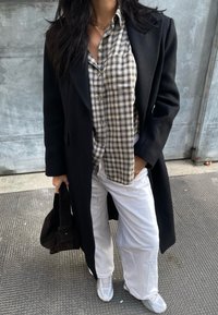 Μαύρο σακάκι πάνω από πουκάμισο σε καρό σε μπεζ και μαύρο, συνδυασμένο με λευκά παντελόνια φαρδιάς γραμμής και γκρι αθλητικά παπούτσια. Μαύρη τσάντα από ύφασμα στο χέρι.