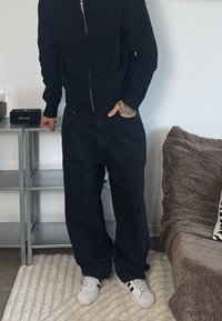 Melns rāvējslēdzēja adīts džemperis, brīva piegriezuma tumši džinsi, balti sporta apavi ar melnām svītrām. Bagātīga tekstūra un tīras līnijas; ikdienišķs apģērbs.