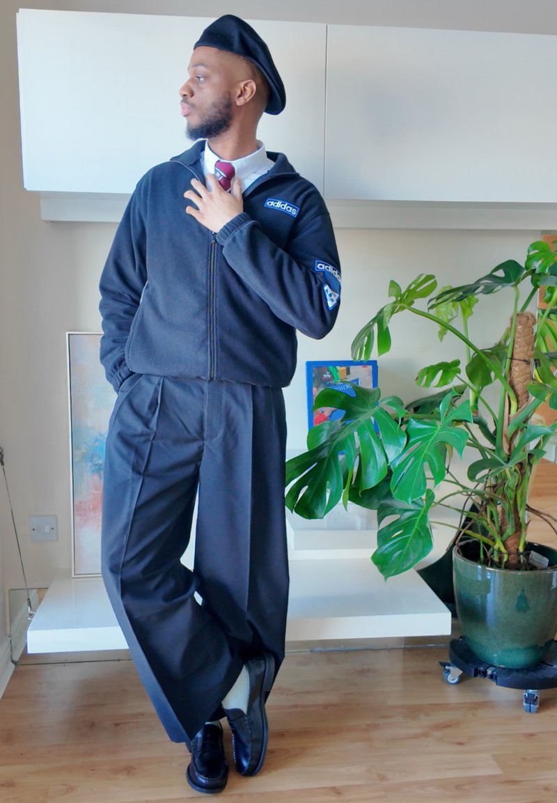 Homme portant une veste Adidas noire, un béret et un pantalon à jambes larges, debout à l'intérieur près d'une grande plante verte en pot sur un sol en bois.