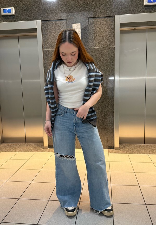 Donna in piedi tra due porte di ascensore che indossa una maglietta con grafica di un cane, un maglione a righe appoggiato sulle spalle, jeans a gamba larga strappati e sneaker.