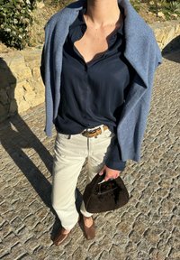 Ναυτικό πουκάμισο με κουμπιά, ανοιχτό μπεζ παντελόνι, καφέ μαλακά παπούτσια, γκρι κασκόλ ριγμένο στους ώμους, κρατώντας μια σκούρα καφέ πλεκτή τσάντα.