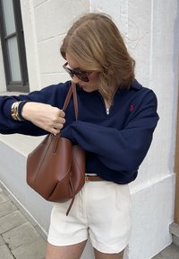 Marinblå zip hoodie, vita shorts, brun läderhandväska, guldfärgad armband. Modellen håller väskan och är klädd i en avslappnad outfit.