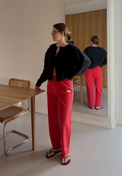 Mujer con suéter negro de felpa, pantalones rojos y sandalias negras de dedo, de pie junto a una mesa de madera con silla de caña en una habitación con pared espejada.