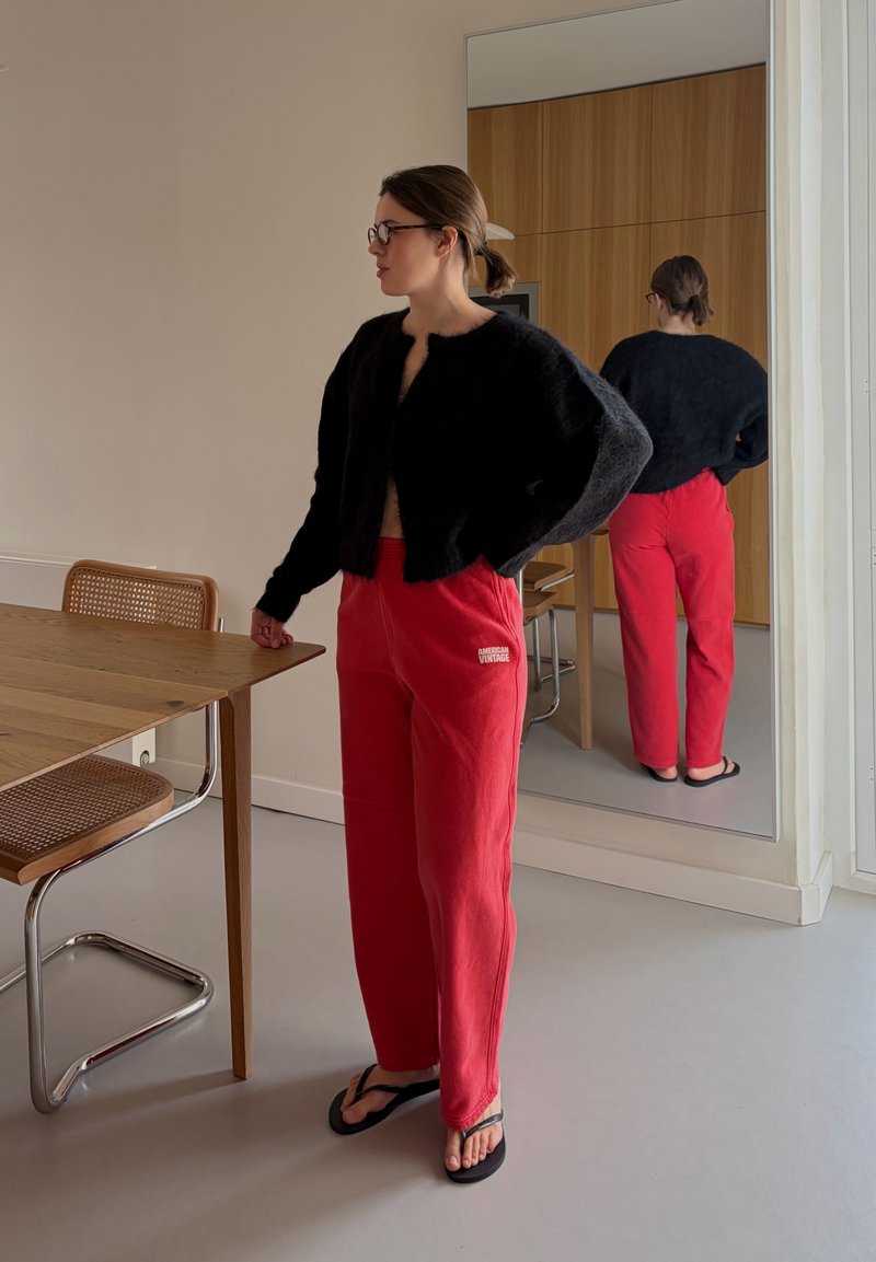 Femme portant un pull noir en tissu pelucheux, un pantalon rouge et des tongs noires, debout près d'une table en bois avec une chaise en osier dans une pièce avec un mur miroir.