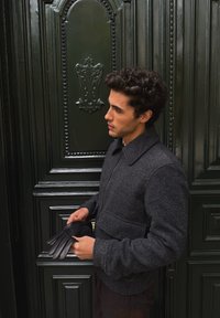 Giacca di lana grigia con cerniera e due tasche, tenuta da una persona con guanti di pelle neri. Porta ornata verde scuro sullo sfondo.