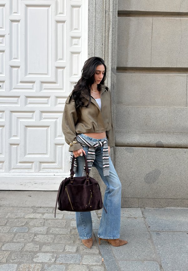 Kvinne med langt mørkt hår iført olivengrønn jakke, blå jeans, brune hæler og stripete genser knyttet i livet, holder en mørk håndveske ved en steinvegg.