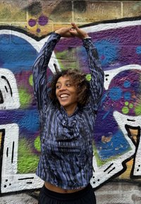 Črn in moder dolg rokav vrhnji del s valovitim vzorcem, ki ima ovratnik in sproščen kroj. Ozadje je obarvane grafitete na opečnati steni.