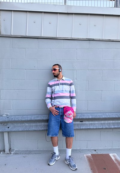 Camisa de manga larga a rayas en azul claro, gris y rosa; pantalones cortos de mezclilla; gorra de camuflaje rosa; zapatillas grises; auriculares; de pie contra una pared gris.