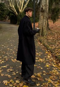 Cappotto lungo nero, guanti neri e scarpe nere. In piedi su un sentiero coperto di foglie autunnali, di profilo con una tazza di caffè in mano.