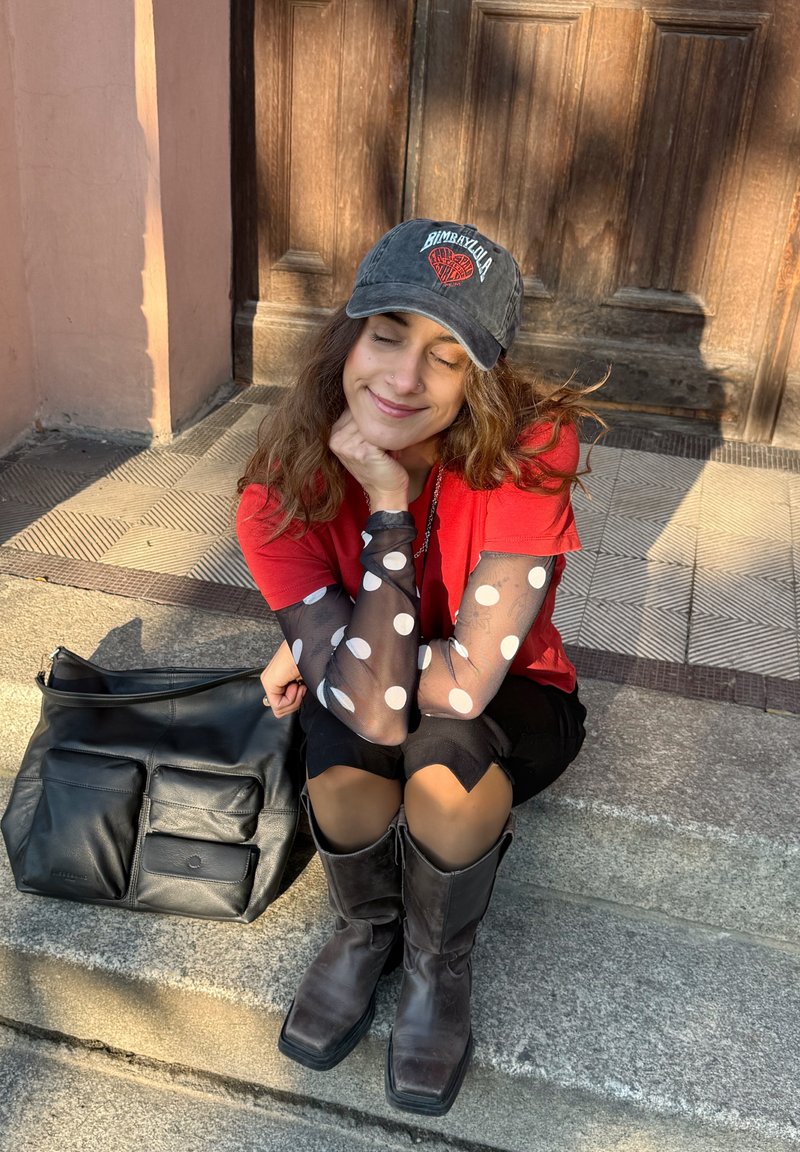 Ung kvinna med slutna ögon, iklädd keps, röd tröja med prickiga nätärmar och svarta stövlar, sittandes på stentrappor med en svart väska bredvid sig.