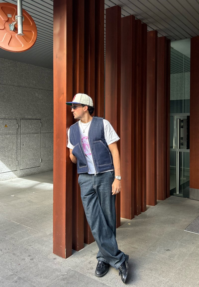 Un gilet en denim sur un t-shirt graphique blanc, un jean baggy foncé, des chaussures noires, des lunettes de soleil et une casquette de couleur claire, adossé à des colonnes en bois.