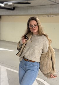 Lysebrun faux pelsjakke over en grå strikke genser, kombinert med lyseblå jeans. Langt hår, briller, minimalt med smykker, står i en parkeringskjeller.