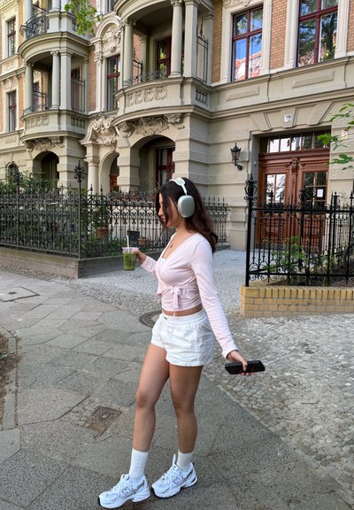 Mujer caminando por un camino de adoquines, vestida con una blusa de manga larga color rosa claro, pantalones cortos blancos, calcetas blancas y auriculares grises, sosteniendo una bebida verde.