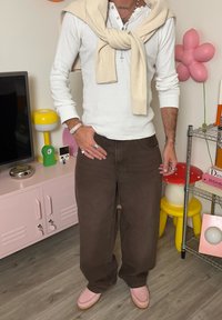 Witte lange mouwen henley, beige trui gedrapeerd, bruine wijde broek, roze instapschoenen. Accessoires zijn ringen en een horloge.