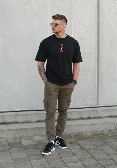 Hombre con gafas de sol y brazo tatuado, vestido con una camiseta negra, pantalones cargo marrones y zapatillas negras, está apoyado contra una pared de hormigón gris.
