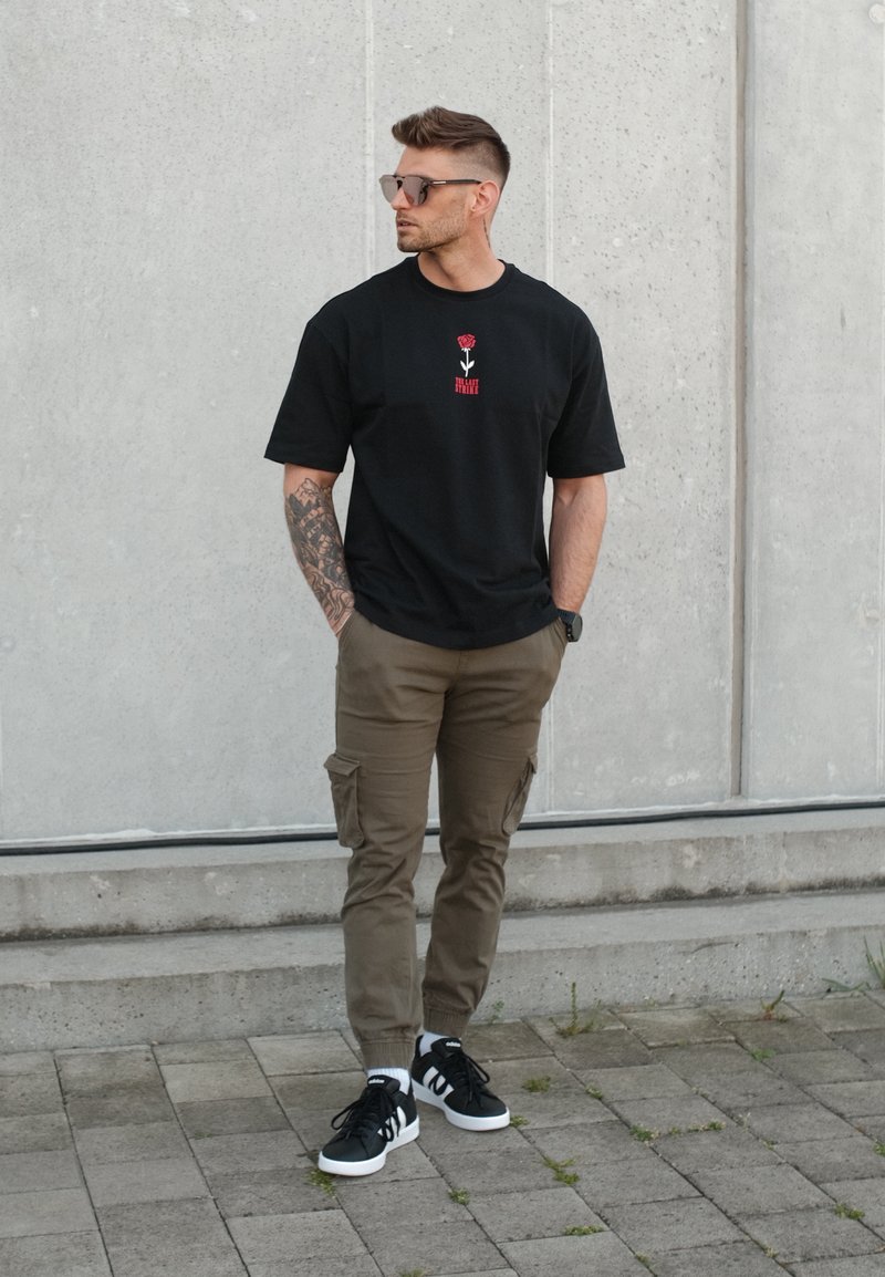 Homme avec des lunettes de soleil et un bras tatoué, portant un t-shirt noir, un pantalon cargo marron et des baskets noires, se tient devant un mur en béton gris.