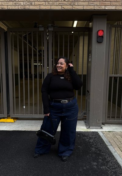 Camiseta negra de manga larga con textura acanalada y logo, pantalones vaqueros de pierna ancha, hebilla de cinturón plateada y bolso negro pequeño, de pie al aire libre cerca de una zona cerrada.