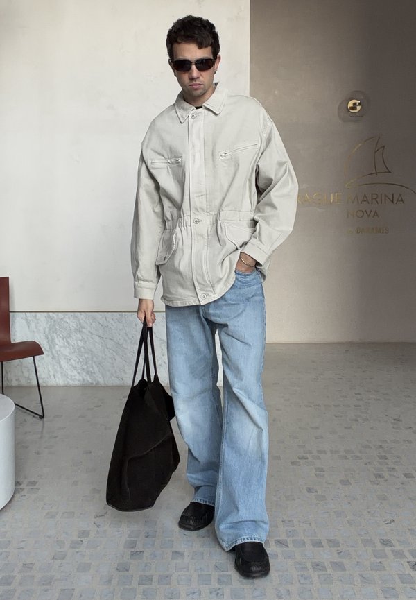 Mężczyzna w okularach przeciwsłonecznych, beżowej kurtce i luźnych niebieskich dżinsach trzyma dużą czarną torbę tote w nowoczesnej przestrzeni wewnętrznej.