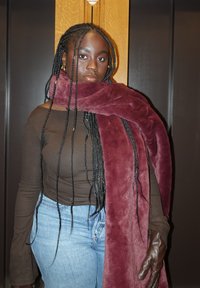 Femme aux longs cheveux tressés portant un haut marron, un jean bleu, une grande écharpe bordeaux et un gant en cuir marron, debout devant un mur en bois et sombre.