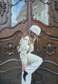 Inimene kreemjas kostüümis ja mustade teravate saapadega, kes kohendab karvast halli mütsi, poseerib kaunilt nikerdatud tumedast puidust ukse lähedal.